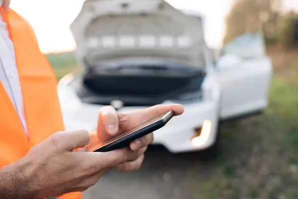 O homem está usando o telefone para pedir ajuda quando seu carro elétrico está quebrado. Um homem de colete de segurança. Um acidente de viação. Ajuda a reparar — Fotografia de Stock