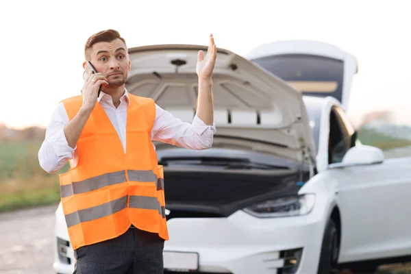 Unge man utanför bilen. En besviken affärsman som pratar i telefon. Bruten bil med öppnat motorhuv. Hjälplös man behöver hjälp och hjälp. Elbilsolycka. Bilproblem. — Stockfoto