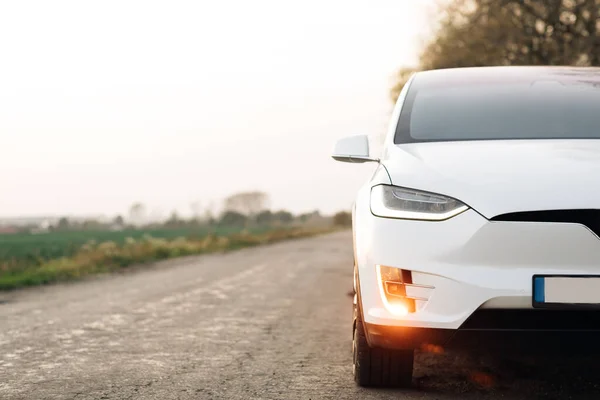 Country Road 'daki elektrikli araba. Ağaçlar ve tarlalar boyunca lüks ve modern bir araç. Çakıl yolunda günbatımında ağaçlı elektrikli araba..