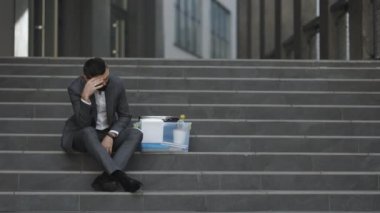 Sağlık maskeli beyaz erkek ofis çalışanı depresyonda bir kutu eşyayla merdivenlerde oturuyor. Endişeli bir konsept. Umutsuzluk içindeki işsiz adam. İşsiz işadamı işini kaybetti