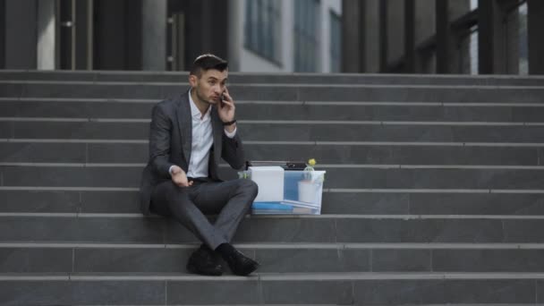 Joven trabajador de oficina sin trabajo con traje sentado en las escaleras al aire libre. Hombre despedido hablando por teléfono con cosas en la caja. Conversación telefónica celular. Hablando por teléfono — Vídeo de stock