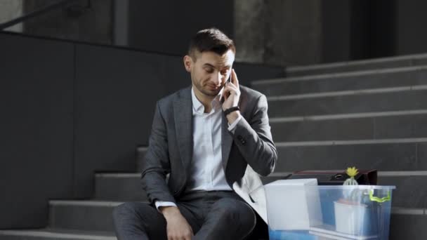 Handsome jobless office worker entrepreneur in formal suit sitting on stairs outdoors. Fired man talking on phone with stuff in box. Cellphone conversation. Speaking on telephone — Wideo stockowe