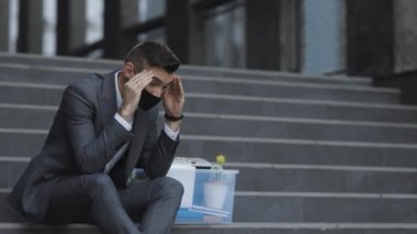 Sağlık maskeli, depresyondaki merdivenlerde bir kutu eşyayla oturan kovulmuş bir ofis çalışanı. İşsiz işadamı işini kaybetti. Endişeli bir konsept. Umutsuzluk içindeki işsiz adam