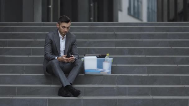 Trauriger Geschäftsmann, der mit einer Kiste voller persönlicher Bürokram auf einer Treppe im Freien sitzt. Verzweifelte Männer verloren ihren Arbeitsplatz und suchten per Smartphone nach einem Job. Pandemische Wirtschaftskrise. Feuriger Mann — Stockvideo