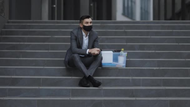 Trauriger junger Geschäftsmann in medizinischer Maske, der auf der Treppe neben dem Bürogebäude sitzt. Ich fühle mich schlecht und ärgere mich. Arbeitsloser Geschäftsmann verlor sein Geschäft. Sorgenvolles Konzept. Arbeitsloser Mann verzweifelt — Stockvideo