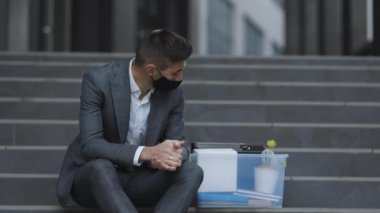 İşsiz işadamı işini kaybetti. Endişeli bir konsept. Umutsuzluk içindeki işsiz adam. Sağlık maskeli kovulmuş erkek ofis çalışanı. Depresyonda, elinde bir kutu eşyayla merdivenlerde oturuyor.