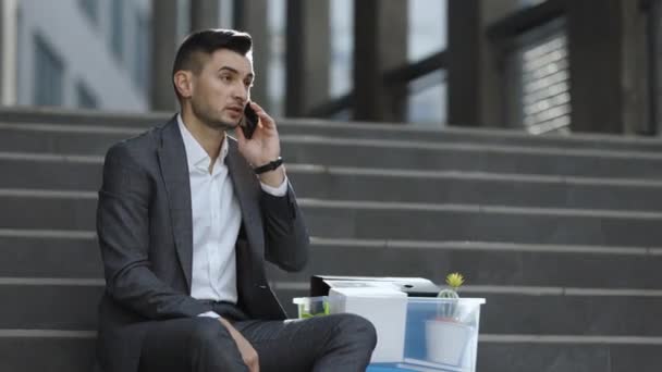 Hombre despedido hablando por teléfono con cosas en la caja. Guapo trabajador de oficina sin trabajo empresario en traje formal sentado en las escaleras al aire libre. Conversación telefónica celular. Hablando por teléfono — Vídeo de stock