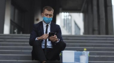 Yoksul erkek iş yerini kaybetti ve akıllı telefondan iş arıyor. Pandemik ekonomik kriz. Tıbbi maskeli kovulmuş bir erkek merdivenlerde kişisel eşyalarıyla oturuyor.
