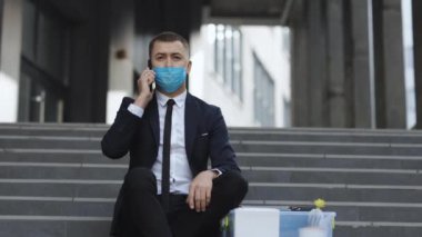 Resmi takım elbiseli, koruyucu maskeli ve merdivenlerde oturan işsiz bir ofis çalışanı. Cep telefonu sohbeti. Telefonda konuşuyorum. Kovulan adam telefonda kutuda eşyalarla konuşuyor.