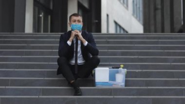 Sağlık maskeli, depresyondaki merdivenlerde bir kutu eşyayla oturan kovulmuş bir ofis çalışanı. Endişeli bir konsept. Umutsuzluk içindeki işsiz adam. İşsiz işadamı işini kaybetti