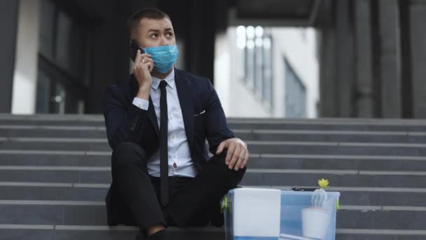 Guapo trabajador de oficina sin trabajo empresario en traje formal y máscara facial protectora sentado en las escaleras. Hombre despedido hablando por teléfono con cosas en la caja. Conversación telefónica celular. Hablando por teléfono — Vídeo de stock