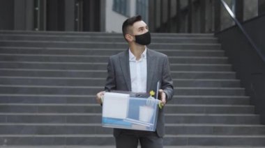 Sağlık maskeli üzgün genç işadamı, kişisel eşyalarıyla birlikte bürodan ayrılan kutuyu taşıyor. İşsizlik parası. İşsizlik yüzünden. Ekonomik kriz.