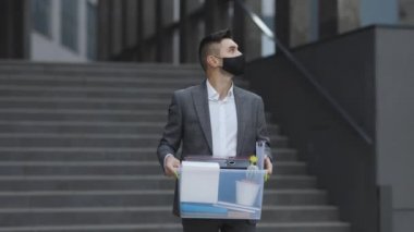 Tıbbi maskeli genç işadamı, ofisten kişisel eşyalarıyla birlikte kutu taşıyor. İşsizlik parası. İşsizlik yüzünden. Ekonomik kriz.