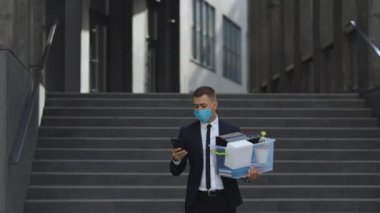 Tıbbi maskeli bir iş adamı. Sokakta yürüyen kişisel eşyalarla dolu bir kutu. Telefon kullanıyor. İş elbisesi. Coronavirus açık havada sosyal uzaklık. Finans ve endüstri. Kovulan adam işini kaybetti.