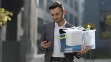 Mutlu iş adamı gülümseyerek ayakta duruyor iş merkezinin yanındaki telefonu kullanıyor. Bir kutu kişisel eşyası olan bir iş adamı telefon mesajlaşması kaydırağını kullanıyor. Portre elbisesi kariyer ofisi yakışıklı teknolojisi