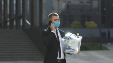 Kovulan iş adamı, modern ofis binasının yakınındaki kutuda eşyalarla telefonda konuşuyor. Koruyucu yüz maskesi takan iş adamı şehir merkezindeki sesli mesajları göndermek için akıllı telefon kullanıyor..