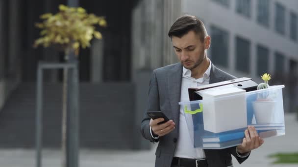 Üzgün genç işadamı ofis binasının yakınındaki telefon mesajlaşmasını kullanıyor. Çaresizlik içindeki erkek ofis çalışanı işini kaybetti. Ciddi teknoloji iletişimi — Stok video