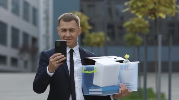 Joven hombre de negocios con caja de cosas personales utiliza mensajes de texto de teléfono de desplazamiento tocando. Feliz hombre de negocios de pie sonriente teléfono de uso cerca del centro de negocios. Traje de retrato carrera oficina masculina tecnología guapo — Vídeo de stock