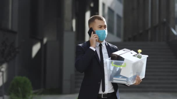 Empresário em máscara protetora usar smartphone para enviar mensagens de voz ao ar livre no centro da cidade. Jovem demitido homem de negócios falando ao telefone com coisas na caixa perto de prédio de escritórios moderno — Vídeo de Stock