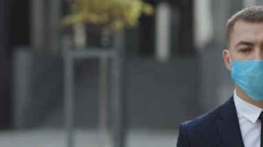 Modern ofis binasının yakınında dikilirken, tıbbi maskeli, üzgün, beyaz, yakışıklı bir adamın yüzünün yarısını kapatın. Koruma maskeli adam sokağa bak.
