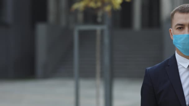 Half face of upset caucasian young handsome man in medical mask looking straight to camera while standing outdoors near modern office building. Man in protection mask look at street — Stock Video