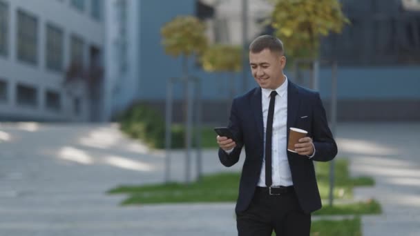 Elegante hombre de negocios que sale de la oficina moderna. Usando su Smartphone. Deslizamiento en la pantalla de móviles. Beber café sabroso. Pareces exitoso, confiado. Celebración de la taza de café en el día de verano — Vídeo de stock