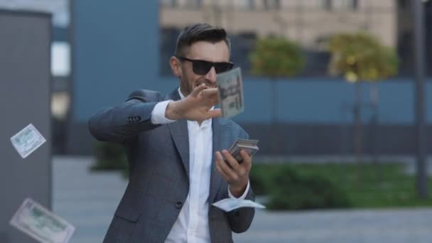 Lluvia de dinero, cayendo dólares. Concepto de negocio exitoso. Empresario exitoso en traje lleva gafas de sol arrojando dinero en la cámara de pie en la calle cerca del edificio de oficinas — Vídeo de stock