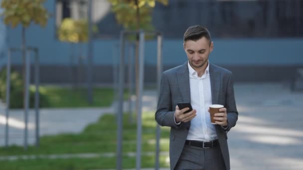 Hombre de negocios barbudo guapo usando teléfono móvil. Hombre joven mensajes de texto, engañando a su teléfono inteligente mientras camina cerca del centro de negocios. Aplicaciones. Negocios. Redes sociales — Vídeo de stock