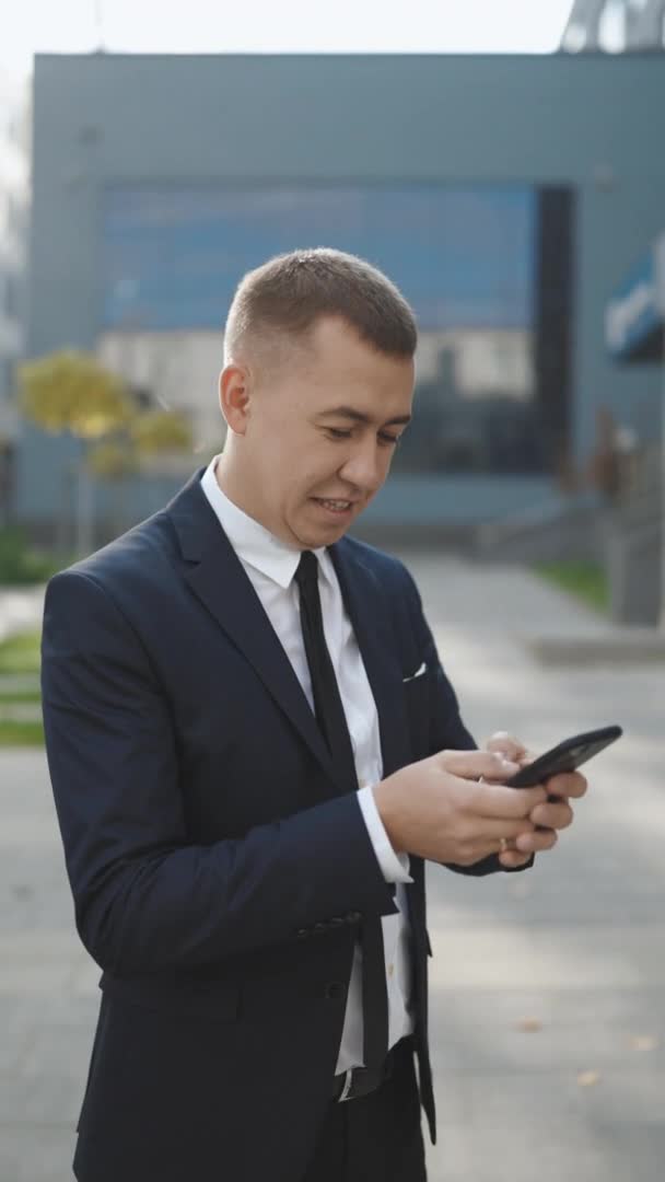 Beau homme d'affaires caucasien tient son smartphone souriant avec charme tout en regardant un contenu vidéo drôle ou des messages texte. Homme réussi en costume élégant en utilisant les applications de médias sociaux — Video