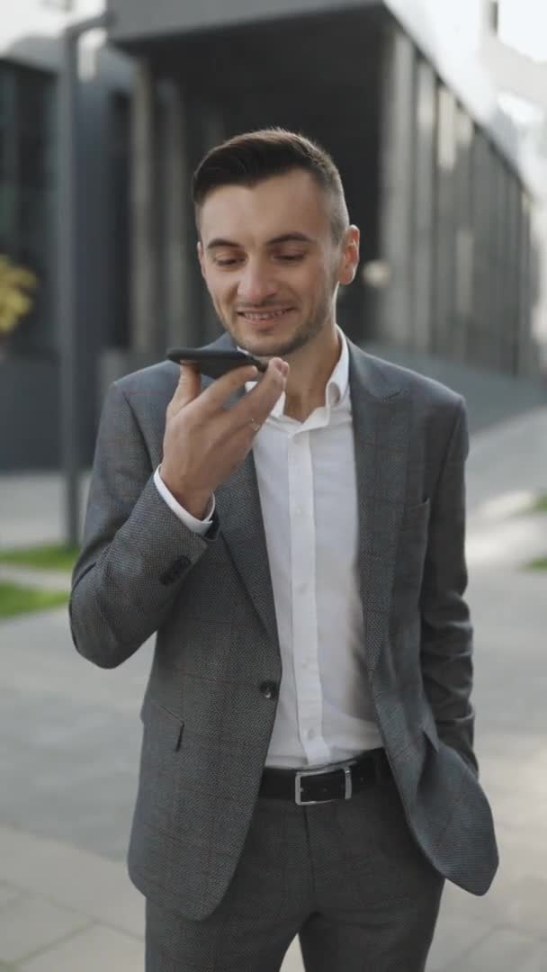 El hombre de negocios utiliza el teléfono inteligente para enviar mensajes de voz al aire libre en el centro de la ciudad. Joven hombre de negocios hablando por teléfono cerca del moderno edificio de oficinas. Vídeo con orientación vertical de la pantalla 9: 16 — Vídeos de Stock