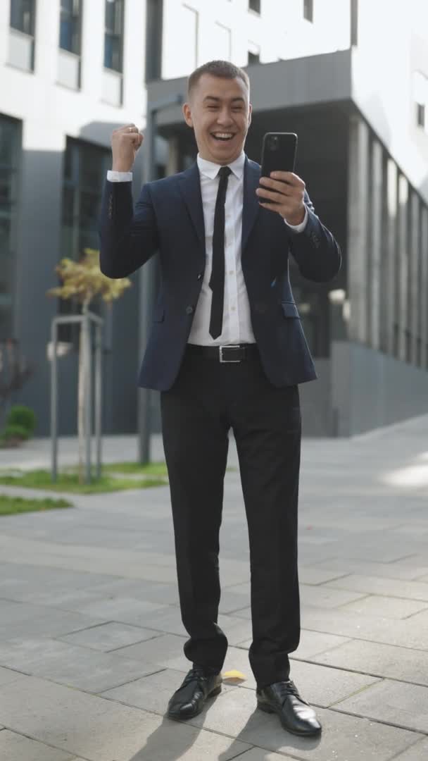 Persona feliz leyendo buenas noticias en el teléfono móvil al aire libre. Un hombre sorprendido celebrando la victoria en un lugar de trabajo remoto. Exitoso en traje jefe empresa confidente. Orientación vertical de la pantalla 9: 16. — Vídeos de Stock