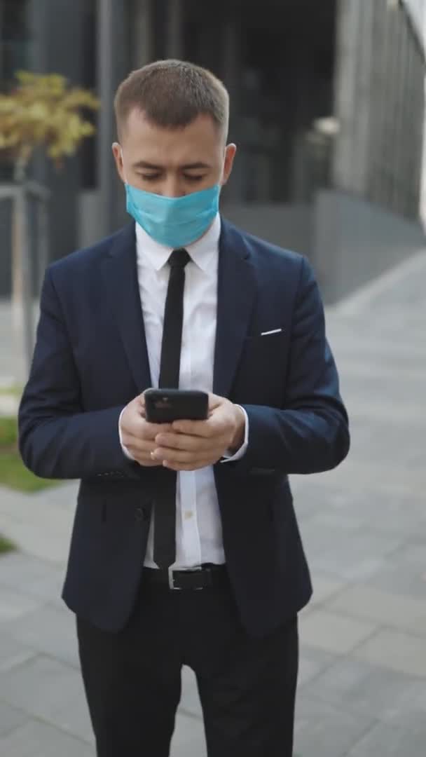 Hombre con mascarilla protectora en la calle mirando la aplicación de redes sociales de navegación de teléfonos inteligentes. Joven pidiendo taxi en aplicación móvil, navegación web escribiendo mensaje. Pantalla de vídeo vertical 9: 16 — Vídeos de Stock