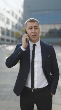 Dışarıda telefonla konuşan yakışıklı bir adamın dikey video portresi. Sokakta akıllı telefondan konuşan bir iş adamı. Mutlu adam dışarıdaki telefonu arıyor. Ofis çalışanı dışarıda konuşuyor.