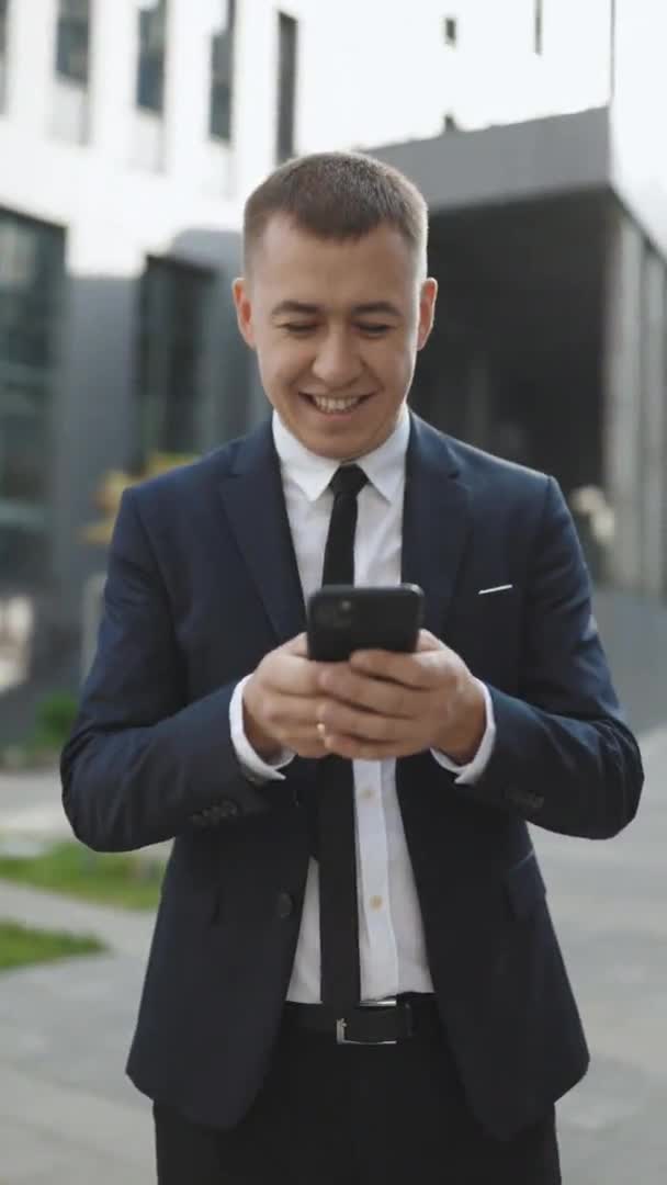 Pantalla de vídeo vertical del hombre sonriendo y utilizando el teléfono móvil sobre el fondo del edificio de oficinas. Tipo europeo guapo sosteniendo y mensajes de texto con el teléfono inteligente. Empresario con tecnología — Vídeos de Stock