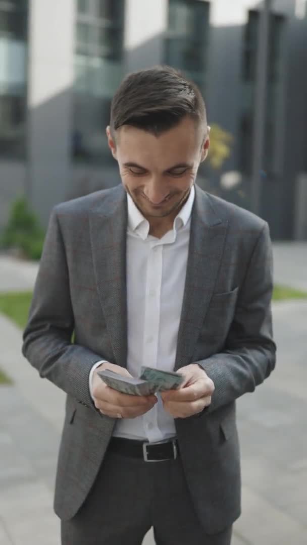 Feliz hombre guapo en estilo de negocio sosteniendo dólares y calculando. Joven guapo rico hombre usando elegante traje contando dinero de pie en la calle cerca de edificio de oficinas vídeo vertical — Vídeos de Stock