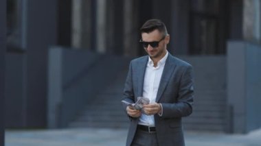 Güneş gözlüklü ve takım elbiseli mutlu bir işadamı para sayıyor ve ofis binasının yanında yürüyor. Başarılı galibiyetini bir sürü dolarla özetliyor. Başarılı görünüyor, kendinden emin.