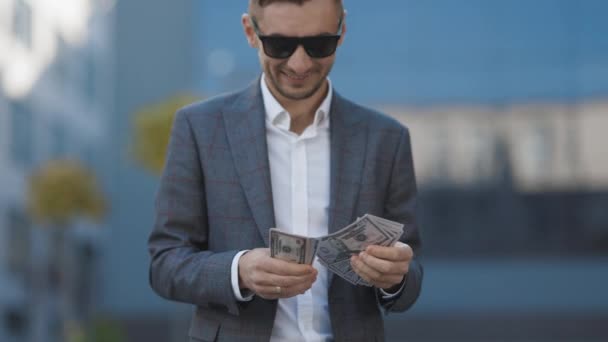 Портрет красивого кавказского молодого бизнесмена в солнечных очках, который считает деньги, стоя на улице. Радостный мужчина с кучей долларов. Роскошная жизнь. Концепция денег Стоковое Видео