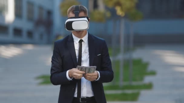 Rik man klädd i virtuell verklighet headset räkna pengar och leende. Ung affärsman som står med dollarpaket nära modern kontorsbyggnad. Rikedom och framgång koncept. 3D VR-glas — Stockvideo