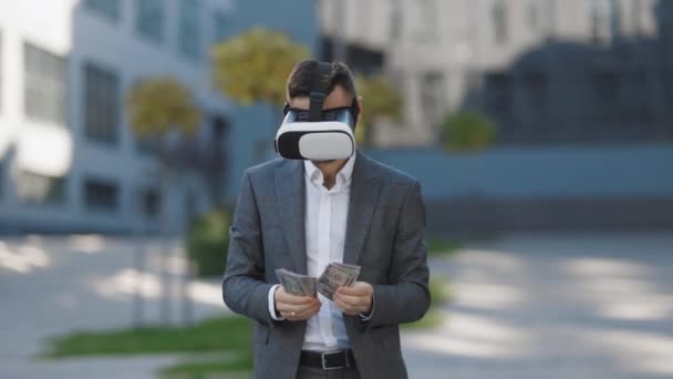 Hombre rico con auriculares de realidad virtual contando dinero. Joven hombre de negocios de pie con paquete de dólares cerca del moderno edificio de oficinas. Concepto de riqueza y éxito. Gafas 3D VR — Vídeo de stock