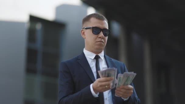 Jonge succesvolle zakenman in een pak dragen een zonnebril tellen geld staan in de buurt van Office Building. Succesvol zakelijk concept. Gelukkig goed uitziende man in zakelijke stijl tellen dollars — Stockvideo