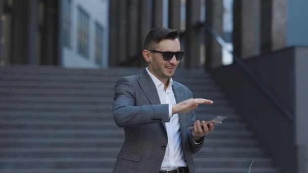 Úspěšný podnikatel v obleku nosí sluneční brýle Hodit peníze do kamery stojící na ulici v blízkosti kancelářské budovy. Prší, padají dolary. Úspěšný podnikatelský koncept — Stock video