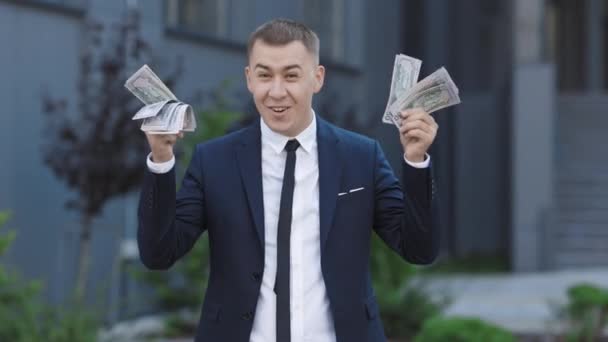 男はカメラを見ながらお金を示して成功を祝う,勝利.屋外だ。お金で幸せな興奮したビジネスマンを驚かせた-米ドル紙幣 — ストック動画