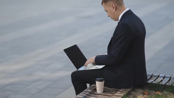 Trabajo a distancia. Un hombre aislado con traje. Un joven hombre de negocios guapo sentado al aire libre trabajando con su portátil. Crisis 2020. Brote de Coronavirus — Vídeos de Stock