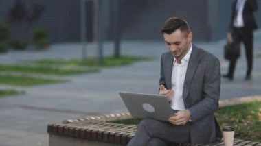 Modern iş adamı dışarıda bankta dizüstü bilgisayarla çalışıyor. Ofisin yanındaki defterde çok güzel konuşan bir video konferansı. Uzak iş yerlerinde jestleri olan erkek profesyonel açıklama projesi.