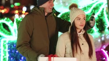 Sevgilisi, karısına Noel hediyesi vererek bayram sürprizi hazırlıyor. Sevgili çift standı. Hediye Kutusu Sarılma Açık Hava 4K