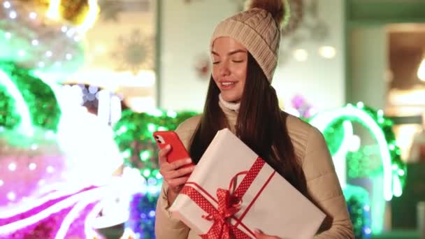 Портрет молодой женщины стоит на улице с рождественским подарком на смартфоне. Подарочная коробка в городе печатает по телефону. Свет вокруг. Улыбаться значит быть счастливым. Подарочная коробка — стоковое видео
