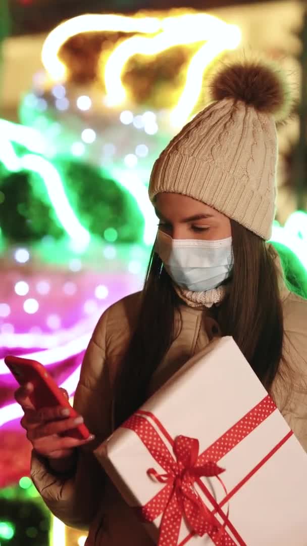 站在街上戴着面具在智能手机上浏览照片的漂亮女性的垂直镜头。带着圣诞礼物的年轻女人在手机上发短信。冬季概念. — 图库视频影像