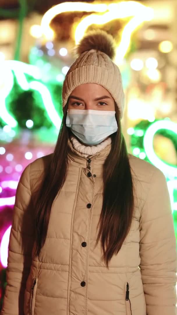 Tiro vertical de primer plano retrato de mujer joven caucásica en máscara facial médica de pie en la ciudad de Navidad decorada. Multitud de personas enfermas covid-19. Coronavirus epidémico. Virus de la corona pandémica de gripe — Vídeos de Stock