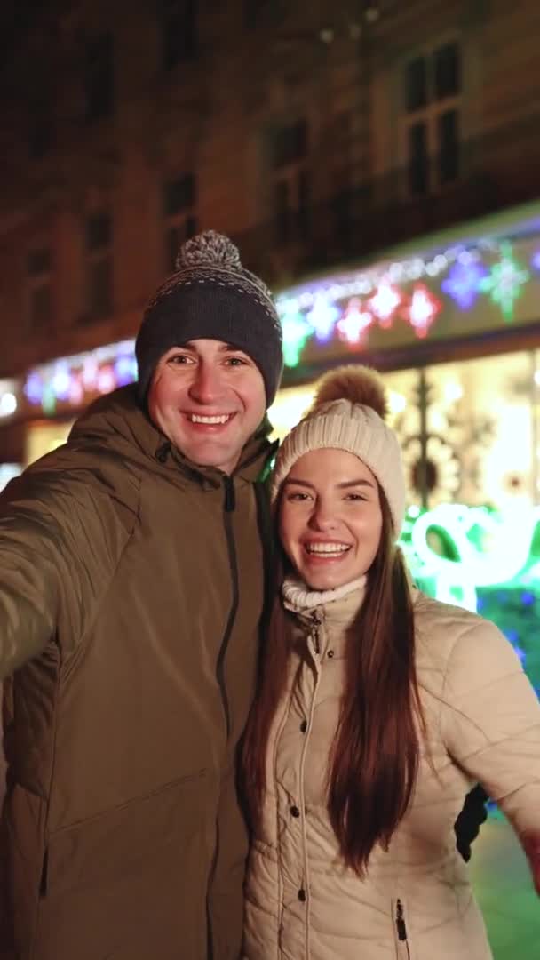 若いカップルの垂直ショットブロガーは、クリスマスの時間に通りでソーシャルページのために話してビデオを作る抱擁立っています.休暇｜Winter Outlook — ストック動画