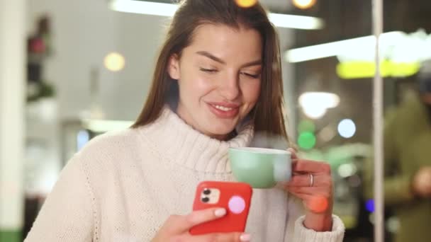 Кавказька весела жінка пробігає по смартфону і п'є зелений чай у затишній кафе. Збереження телефонного пристрою з сенсорним екраном за допомогою інтернет технологій перегляду, пошук гаджета — стокове відео